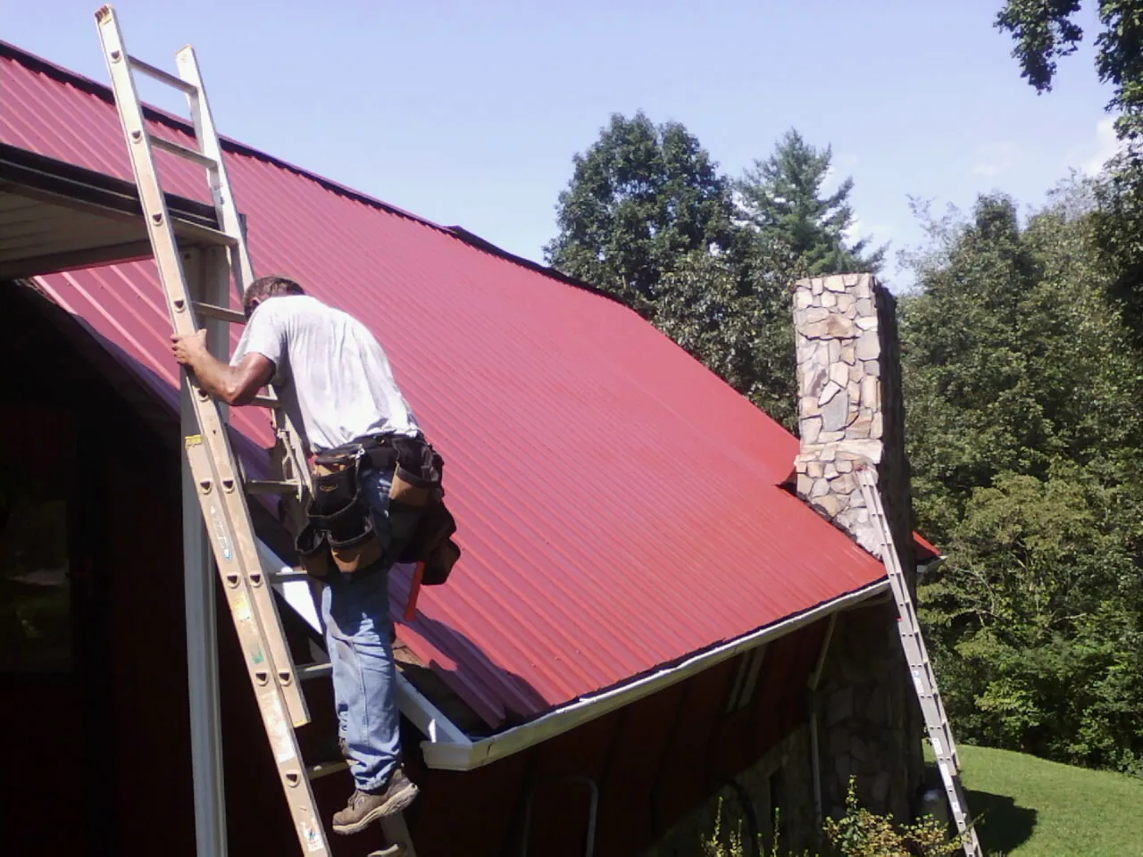 Roofer on a ladder beside a red metal roof and stone chimney — Soffit & Fascia Repair by Roofing Brothers in Toddville